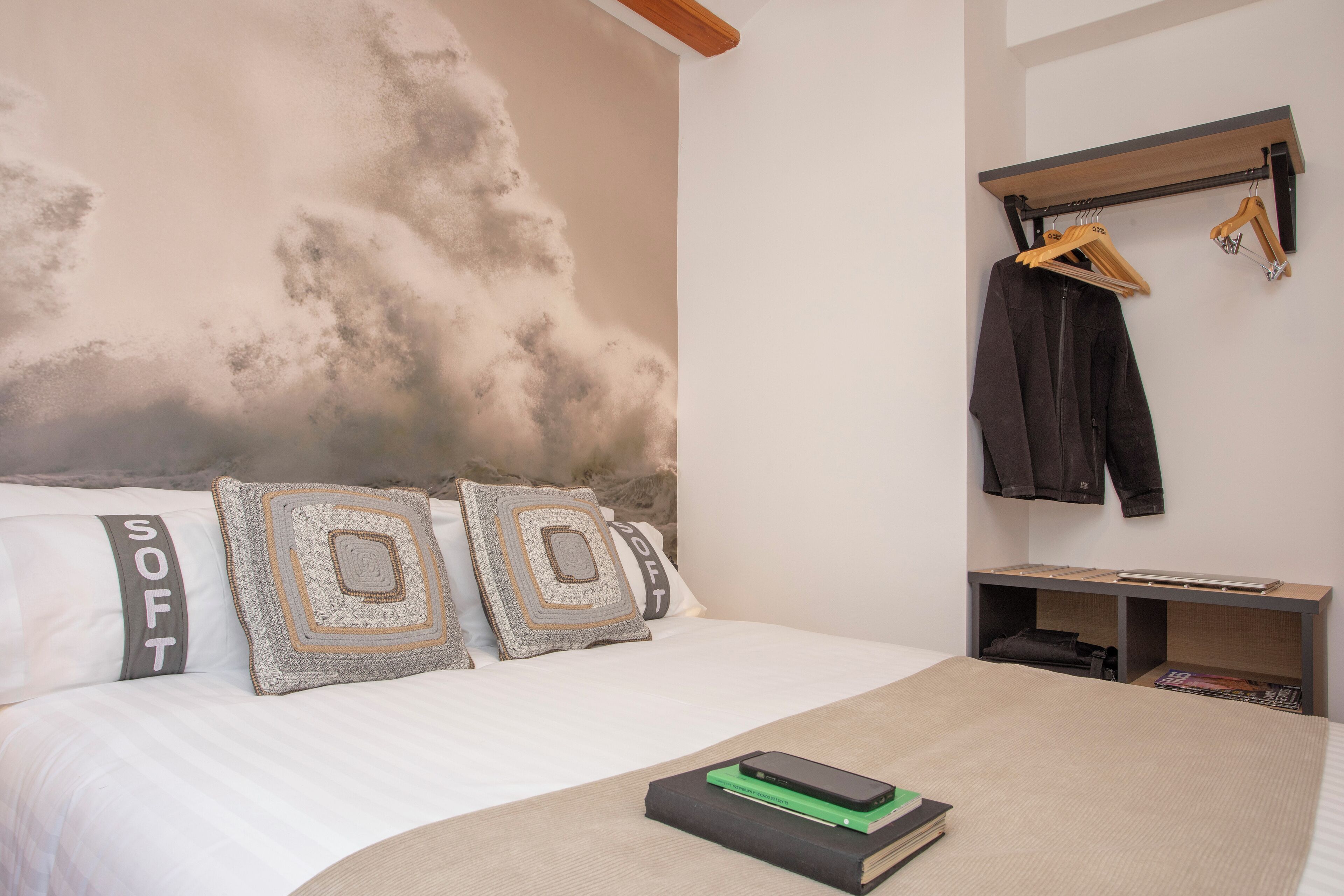 A bedroom with a bed, pillows, a bedside table, a hanging coat rack, and a wall with a cloud mural.
