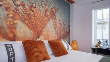 A bedroom with a large bed, two orange pillows, a white pillow with "SOFT" text, a wall mural, a window with a view, and a small table with a lamp.