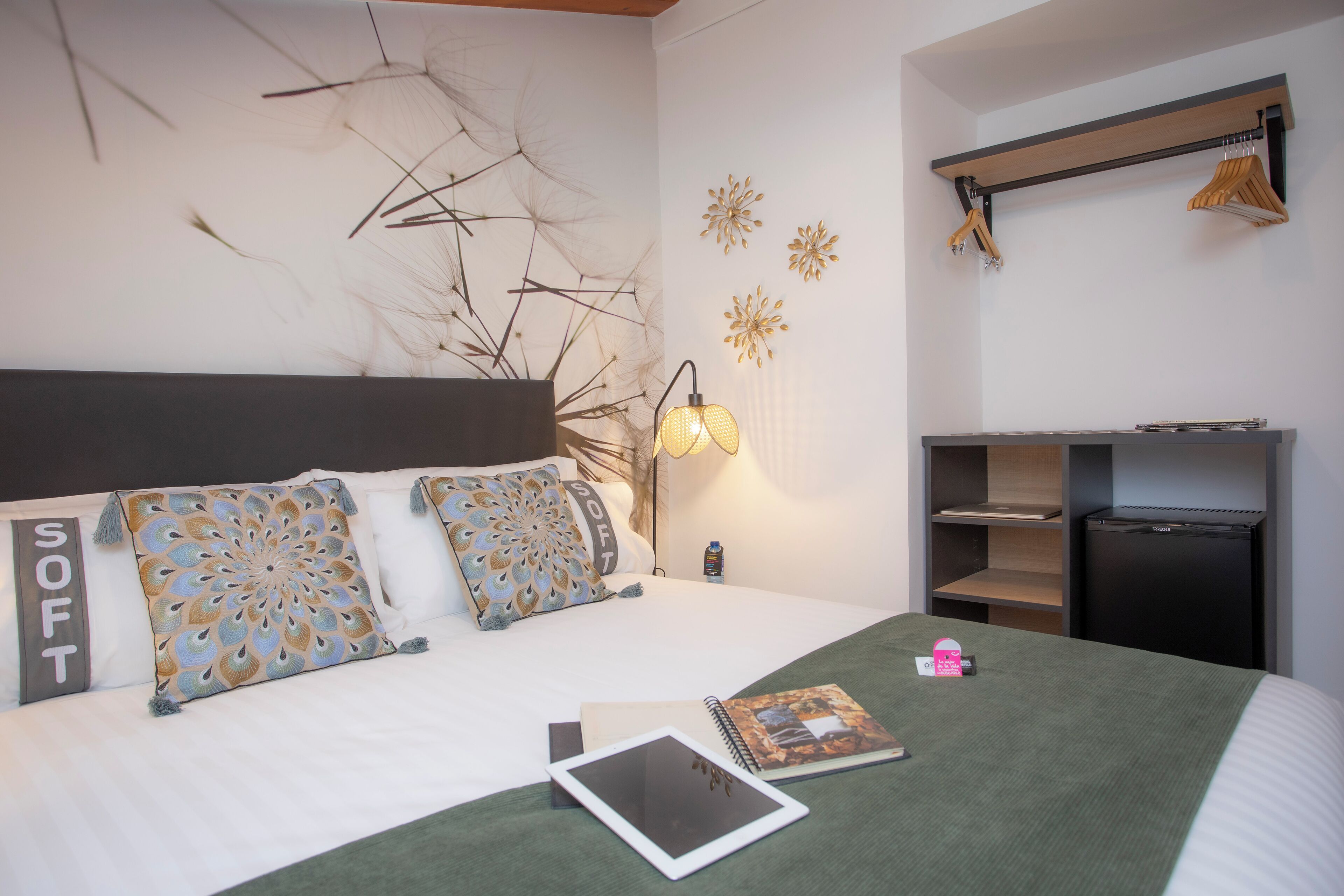 Een netjes opgemaakt bed met gestreepte kussens, een bedlampje, een klein bureau met een computer en een plank met kledinghangers.