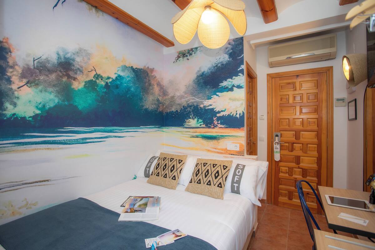 Une chambre avec une grande fresque représentant une scène de plage, une porte en bois, un lit avec des coussins à motifs et une petite table avec une chaise.