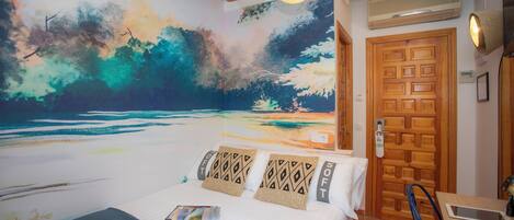 Un dormitorio con un amplio mural de una escena de playa, una puerta de madera, una cama con almohadones estampados y una pequeña mesa con una silla.