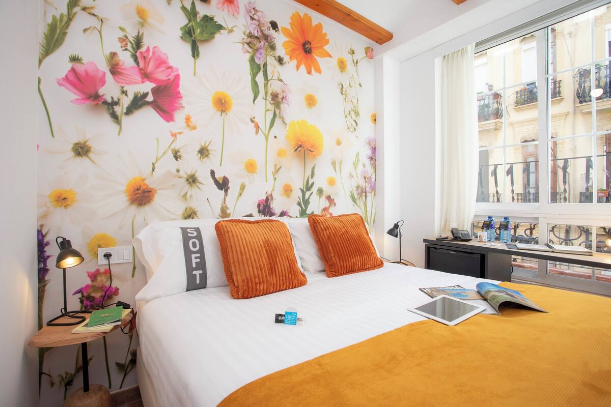 Une chambre d’hôtel avec un grand lit, deux oreillers orange, une table de chevet avec une lampe, un bureau avec un ordinateur portable et une fenêtre donnant sur des immeubles.