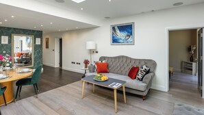 A spacious living room with a grey sofa, a dining table with chairs, a coffee table with fruit, and a painting on the wall. - Apartment in Guildford with Parking (Guildford)