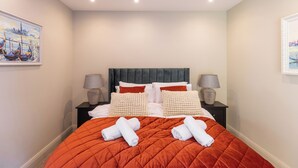 A neatly made bed with a red quilt, two white towels, and a grey upholstered headboard. There are two bedside lamps on black nightstands, and a framed painting on the wall. - Apartment in Guildford with Parking (Guildford)