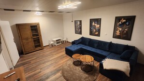 A modern living room with a blue sectional sofa, a wooden coffee table, and a small round table. There is a white dining table with chairs, a wooden cabinet with glass doors, and three framed floral paintings on the wall. - Large family vacation apartment (100 m²) near SPO - dogs welcome 1000m2 garden (Poppenbüll)