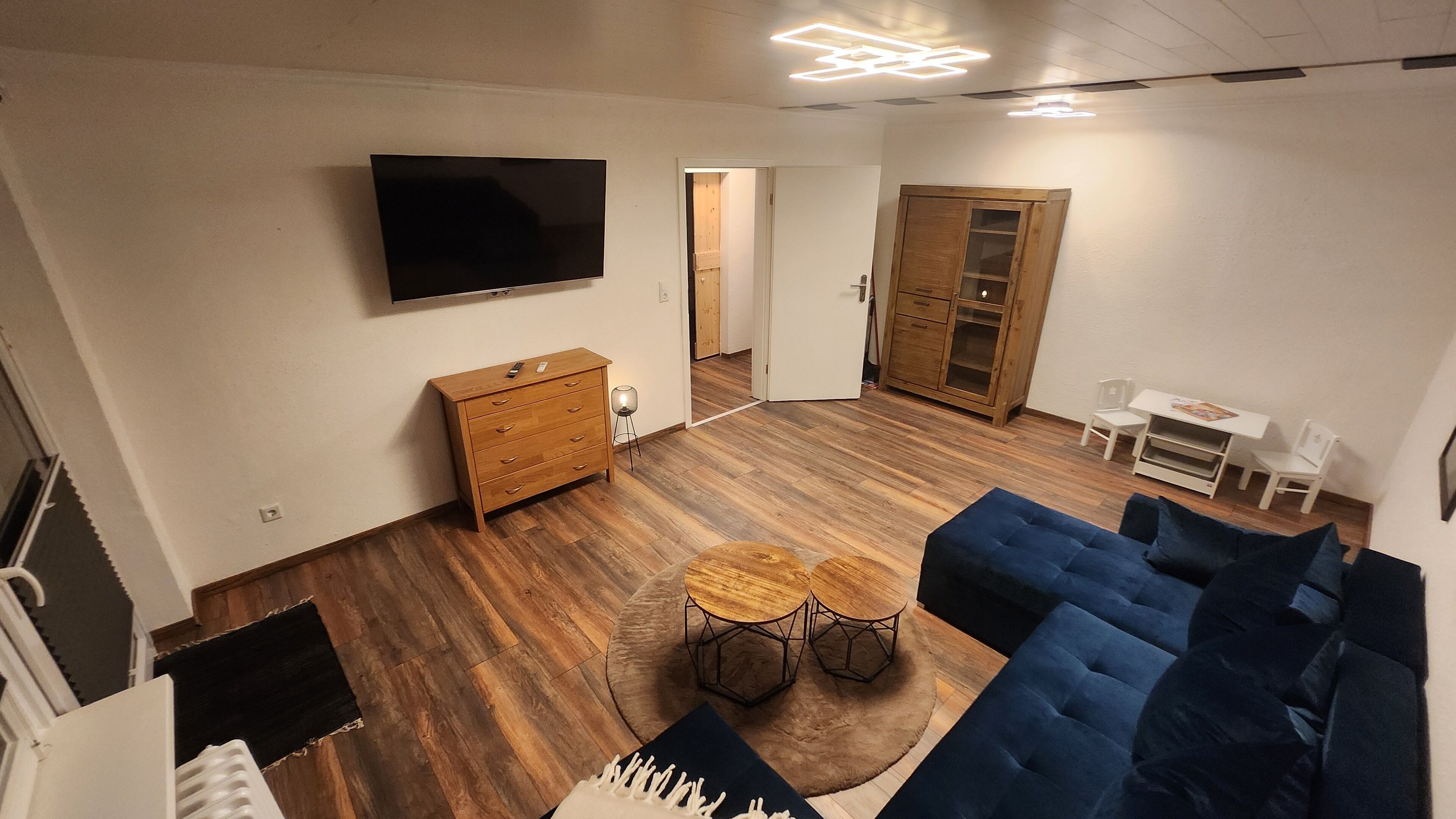 Ein modernes Wohnzimmer mit einem blauen Sofa, Holzböden, einem an der Wand befestigten Fernseher, einem Holzkasten und einer kleinen Essecke mit Tisch und Stühlen.
