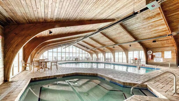 Une piscine intérieure avec des poutres en bois et un sol en briques, entourée de tables et de chaises.