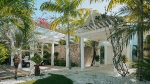 Un espacio al aire libre techado, con pared de piedra, pérgola blanca y diversas plantas tropicales.