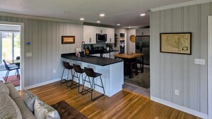 A modern kitchen with a central island, bar stools, and a dining area with a wooden table and chairs. - View With a Room │ Mountain Home in Wine Country (Afton)