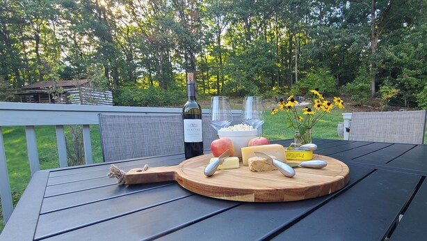 A wooden cutting board with cheese, apples, and a bottle of wine on an outdoor table. - View With a Room │ Mountain Home in Wine Country (Afton)