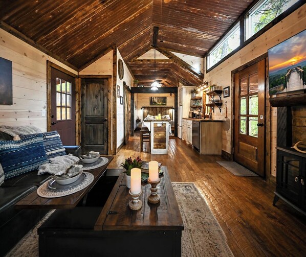 A cozy cabin interior with a wooden ceiling, a dining area, a kitchen, and a living room with a sofa and a fireplace.