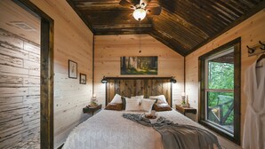 Een slaapkamer in een houten hutje, met een bed, kussens, een schilderij, een raam en een plafondventilator.