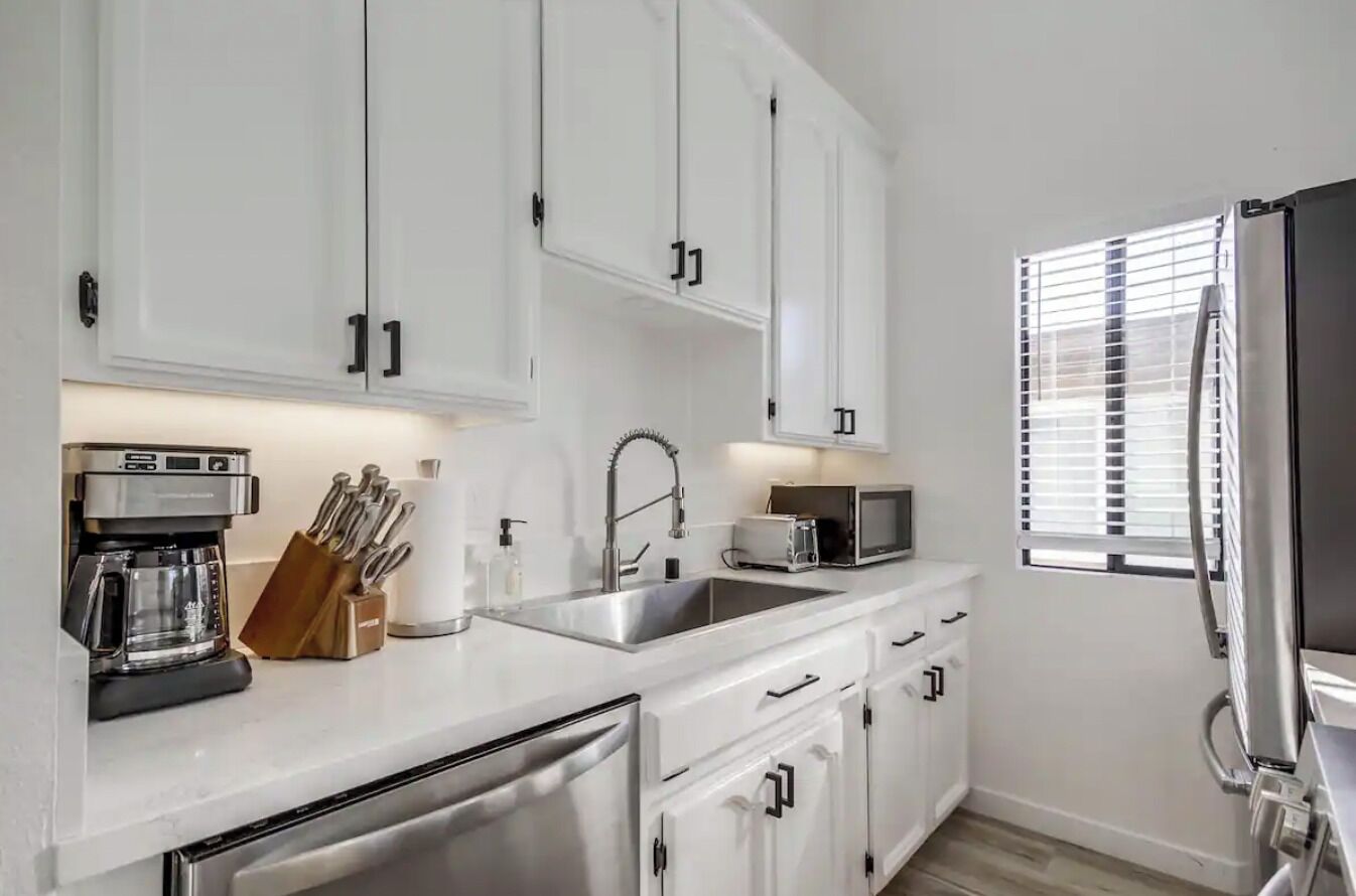 A modern kitchen with white cabinets, stainless steel appliances, and a window with blinds.