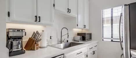 Une cuisine moderne avec des meubles blancs, des appareils en acier inoxydable et une fenêtre équipée de stores.