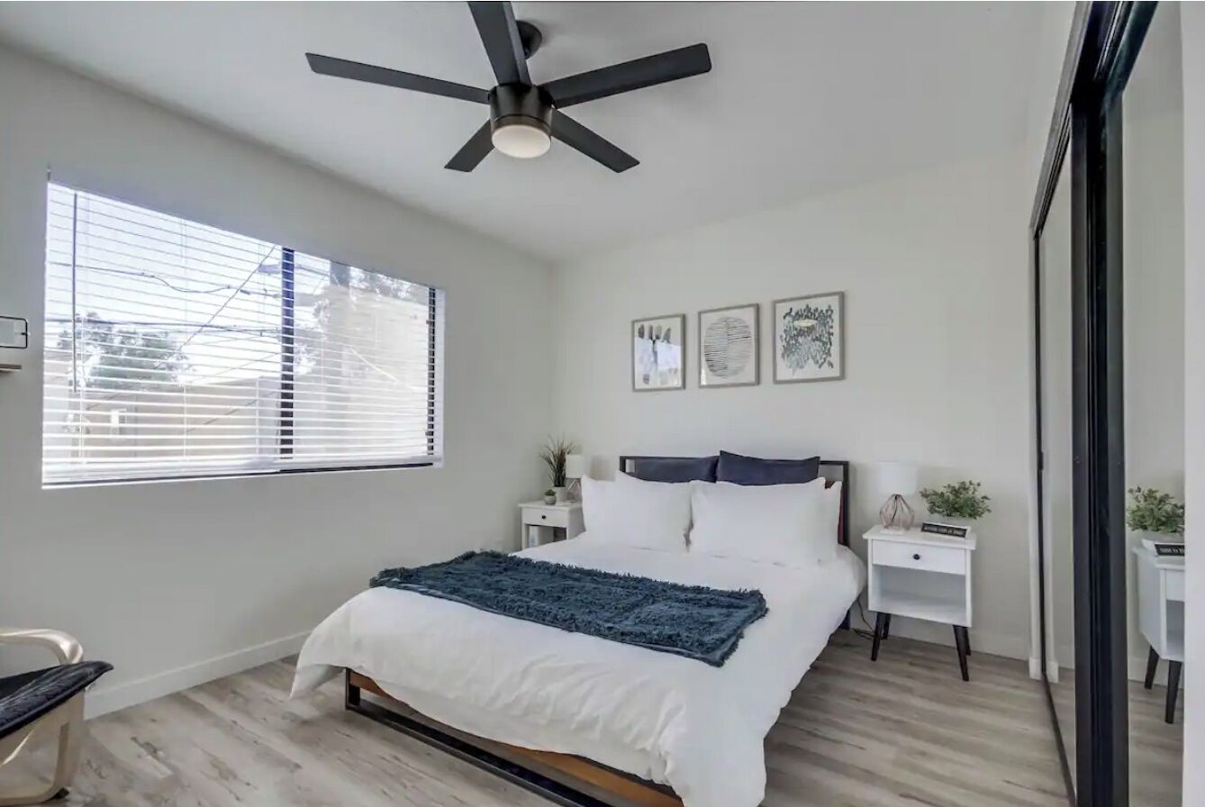 A bedroom with a bed, a ceiling fan, a window with blinds, bedside tables with lamps, and framed artwork on the wall.