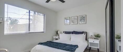A bedroom with a bed, a ceiling fan, a window with blinds, bedside tables with lamps, and framed artwork on the wall.