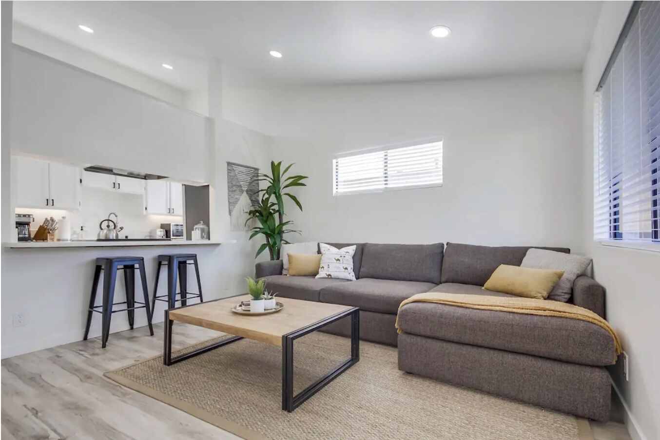 Una sala moderna con un sofá gris, dos taburetes negros, una mesa de centro de madera y una cocina con armarios blancos y una planta.