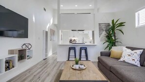 Un soggiorno moderno con TV a schermo piatto, un tavolino con piante, un divano grigio con cuscini decorati e un angolo cucina con isola centrale e sgabelli da bar.