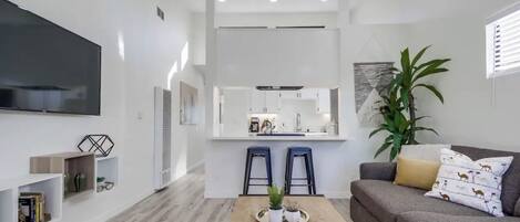 A modern living room with a flat-screen TV, a coffee table with plants, a grey sofa with patterned cushions, and a kitchen area with a central island and bar stools.
