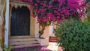 Un bâtiment doté d’une entrée arquée de style traditionnel, d’un escalier menant à l’entrée et d’un jardin agrémenté de fleurs violettes.