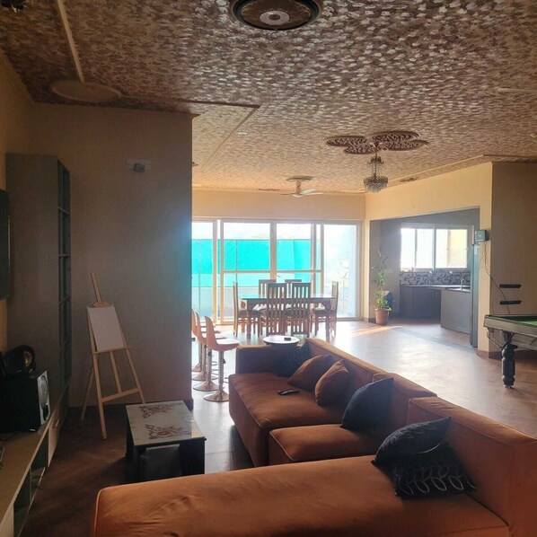 A spacious living area with a large sofa, a dining table, and chairs. There is a ceiling fan, a ceiling light fixture, and a view of the outside through large windows.