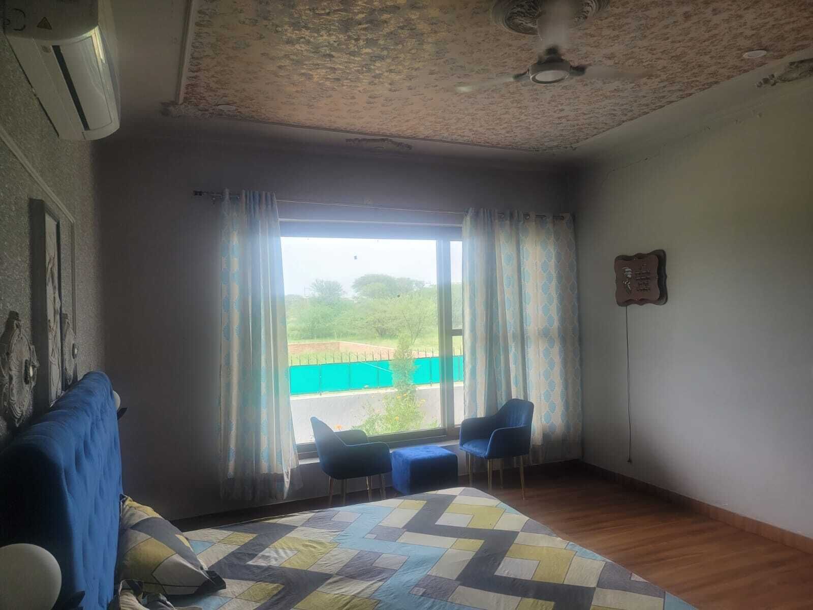 Une pièce comprenant un lit, un bureau, deux chaises, une fenêtre avec des rideaux et un ventilateur de plafond.