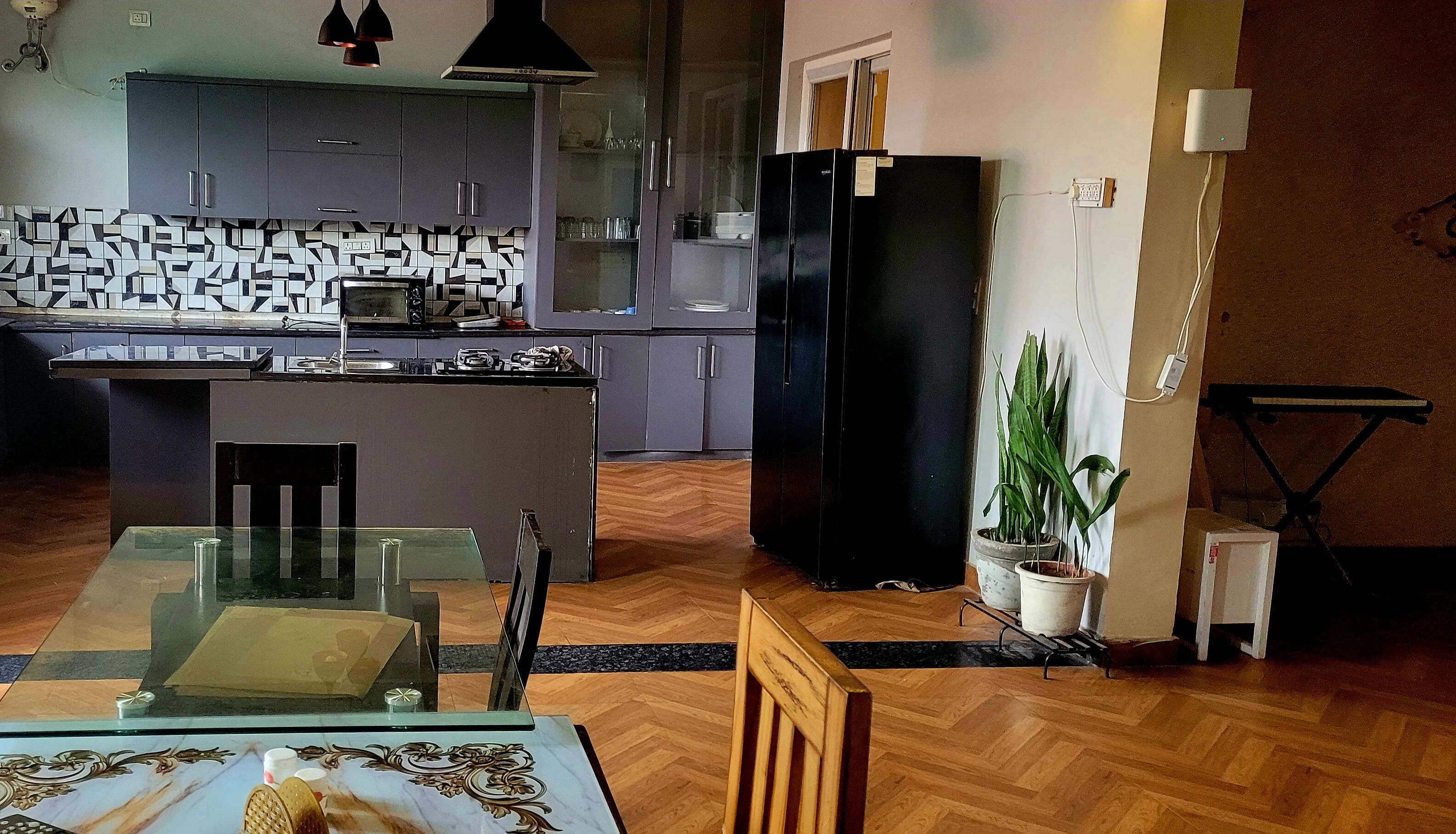 Une cuisine moderne avec des meubles gris, un réfrigérateur noir et un coin repas comprenant une table et des chaises en verre.