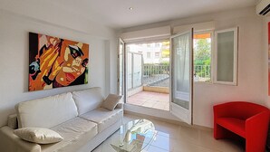 A modern living room with a glass coffee table, a beige sofa, a red armchair, and a large abstract painting on the wall. - HENRI CAMILLE AGENCY - Bosquets - Studio w/ terrace close center (Cannes)