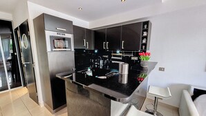A modern kitchen with black cabinets, a stainless steel refrigerator, and a small island with a sink. There is a vase with red flowers on the island. - HENRI CAMILLE AGENCY - Bosquets - Studio w/ terrace close center (Cannes)