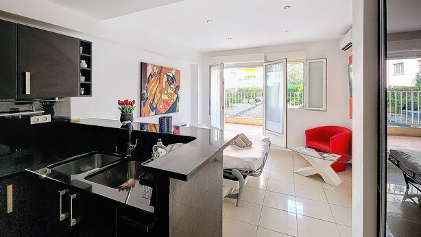 A modern kitchen with a black countertop and stainless steel sink, a dining area with a red chair, and a balcony with a view of greenery. - HENRI CAMILLE AGENCY - Bosquets - Studio w/ terrace close center (Cannes)