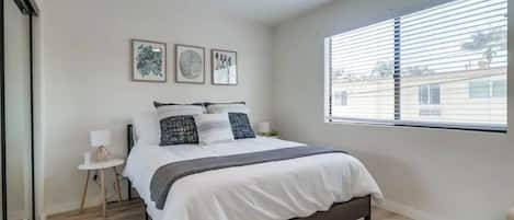 Un dormitorio con cama, ventilador de techo, ventana con persianas y una mesita de noche pequeña con lámpara.