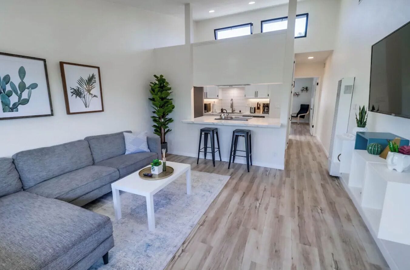 Una sala de estar moderna con un sofá gris, una mesa de centro blanca y una cocina con isla central y taburetes de bar.
