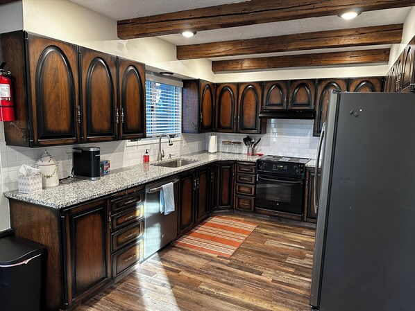 A kitchen with dark wooden cabinets, a granite countertop, a stainless steel sink, a black refrigerator, and a wooden floor. - Beautiful & Spacious 7-Beds by 𝐁𝐘𝐔𝐈 (Rexburg)