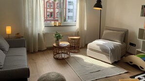 Un salon chaleureux avec un canapé, une petite table sur laquelle est posée une plante, un lampadaire et une vue sur un bâtiment rouge, visible à travers une fenêtre.