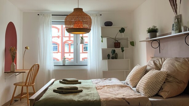 Une chambre à coucher comprenant un lit, une chaise, un bureau et une fenêtre donnant sur des immeubles.