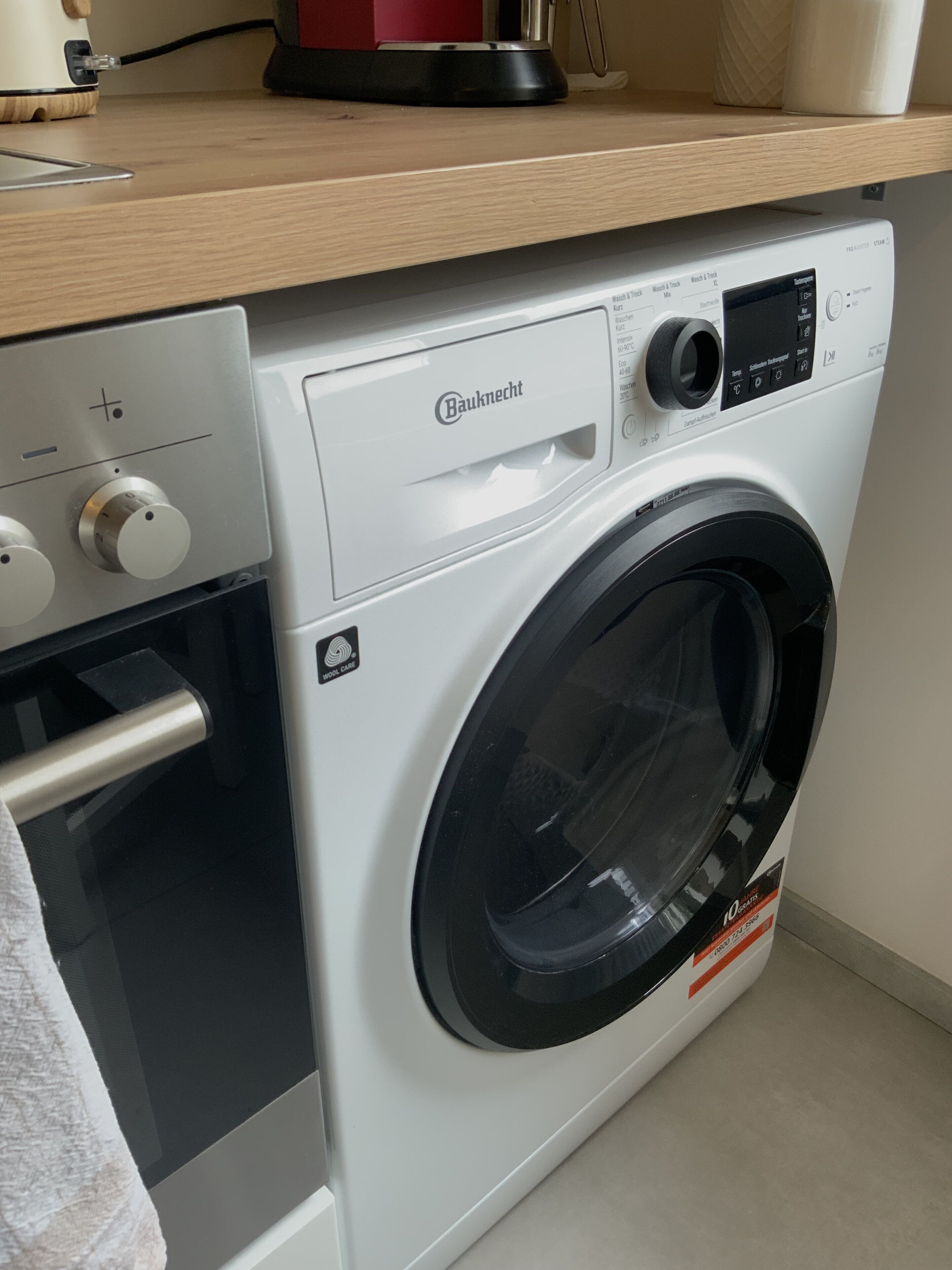 Eine Bauknecht Waschmaschine mit Frontlader, ein Bedienfeld, eine Holz Arbeitsplatte darüber und eine weiße Wand im Hintergrund.