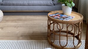 Un salon avec un canapé gris, une table ronde en osier sur laquelle sont posés des magazines, une plante en pot et des éléments de décoration murale.
