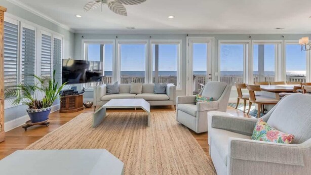 A spacious living room with a large TV, comfortable seating, a ceiling fan, and a view of the ocean through large windows. - 6br Oceanfront Cottage w/ awesome views and short walk to the boardwalk. (Carolina Beach)