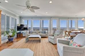 Un salón amplio con un televisor grande, asientos cómodos, un ventilador de techo y vista al océano a través de amplias ventanas.
