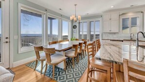 A spacious dining area with a large table and chairs, a kitchen with white cabinets, and large windows offering a view. - 6br Oceanfront Cottage w/ awesome views and short walk to the boardwalk. (Carolina Beach)