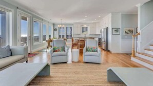 A spacious living area with a sofa, two armchairs, a coffee table, and a dining table with chairs. There is a kitchen area with cabinets and a refrigerator in the background. - 6br Oceanfront Cottage w/ awesome views and short walk to the boardwalk. (Carolina Beach)