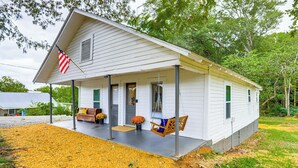 A white single-story house with a porch, a swing chair, and a small garden with orange flowers. - Downtown Heflin Cottage w/ Deck & Fire Pit! (Heflin)