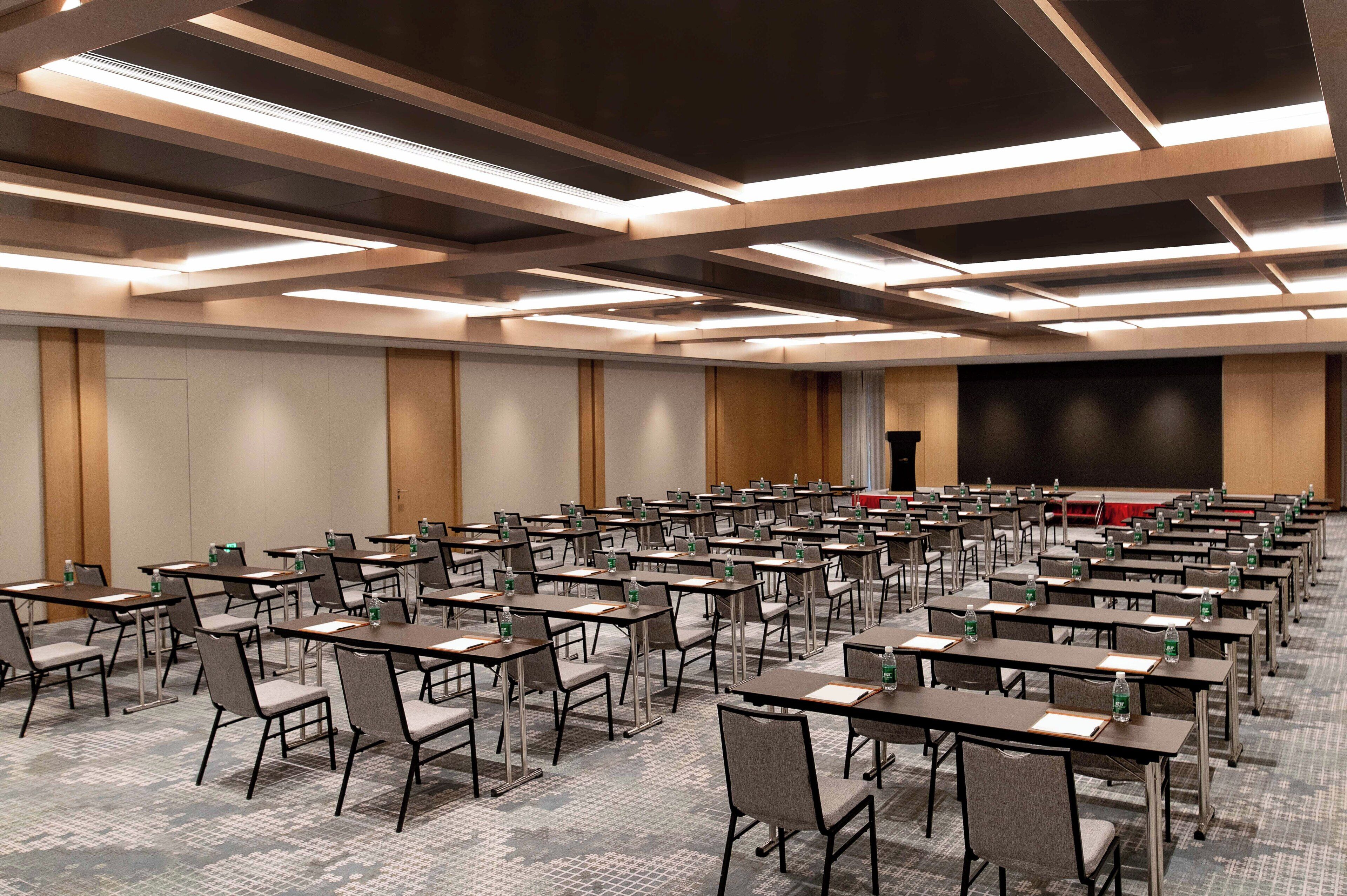 A conference room with rows of tables and chairs, equipped with water bottles, and a large screen at the front.