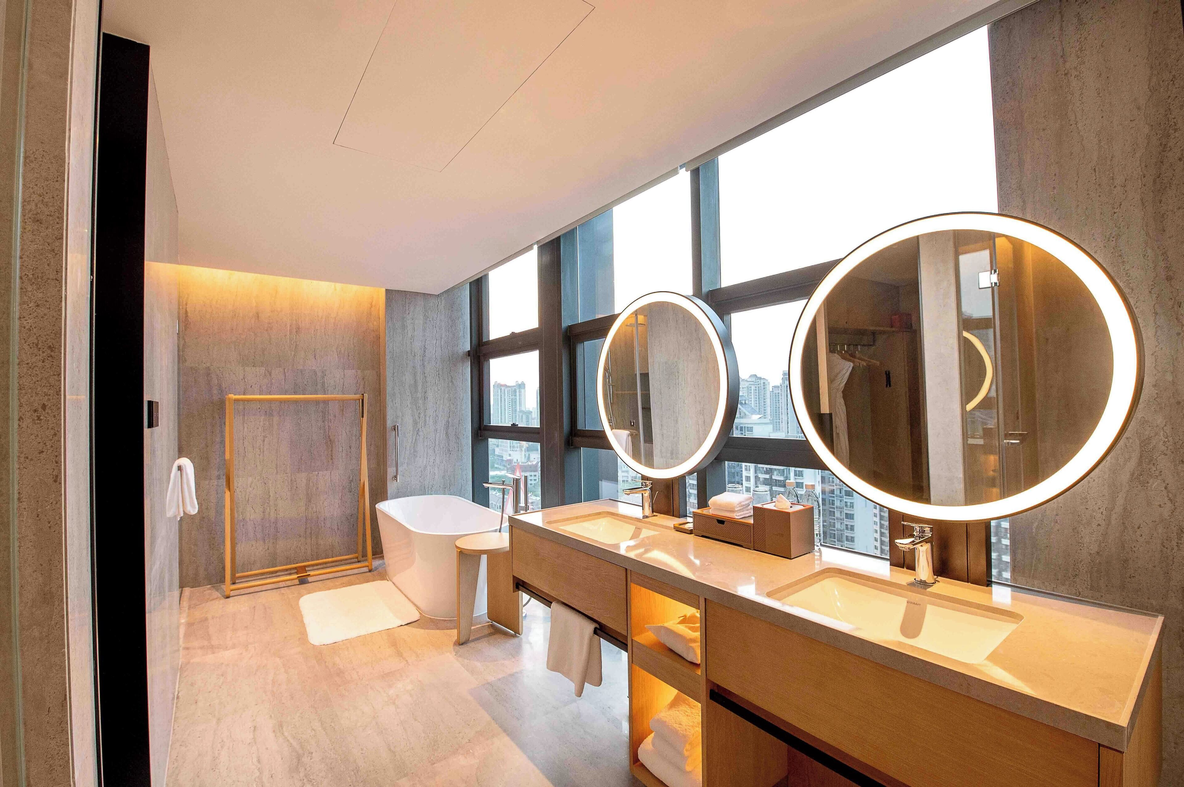 Un baño moderno con una bañera independiente, un tocador con un espejo grande y vistas a la ciudad a través de amplias ventanas.