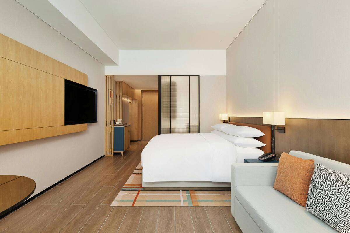Ein modernes Hotelzimmer mit einem großen Bett, einem an der Wand montierten Fernseher, einem Nachttisch mit Lampe und einer Couch mit Kissen.
