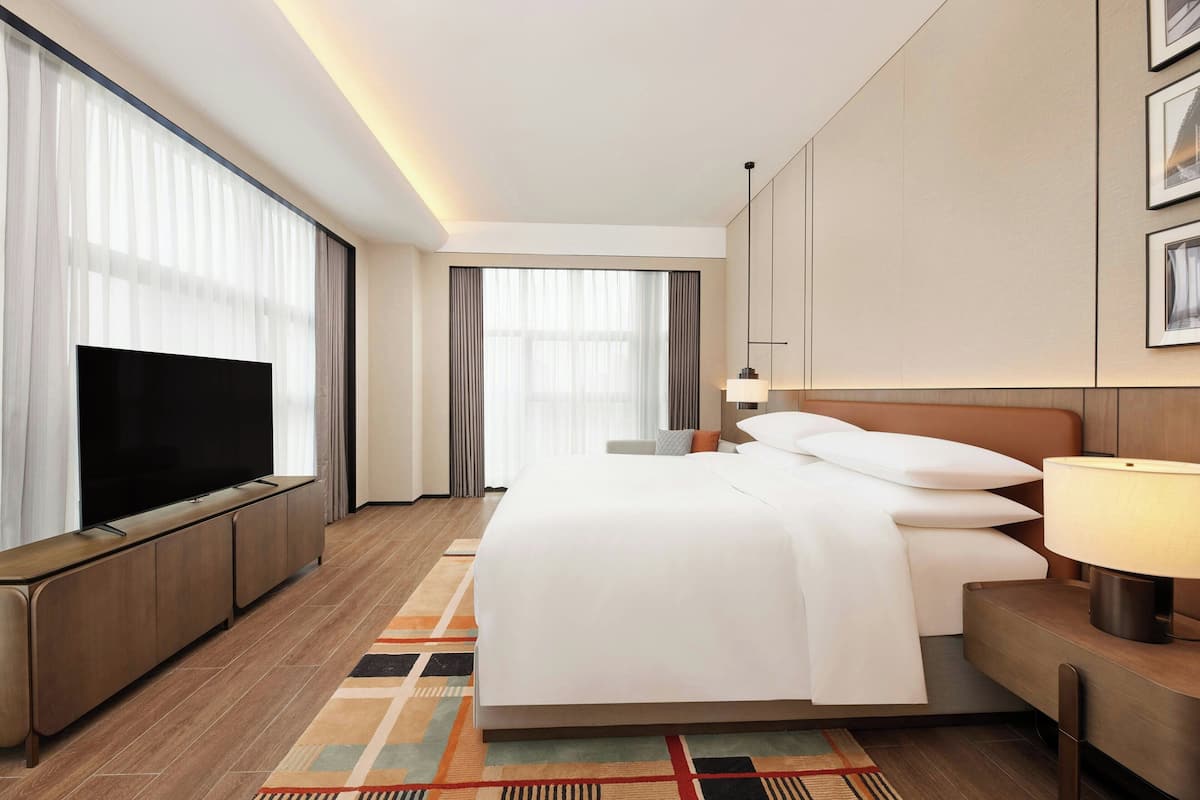 Ein Hotelzimmer mit einem großen Flachbildfernseher, einem ordentlich bezogenen Bett, einem Nachttisch mit Lampe und einem großen Fenster mit Vorhängen.
