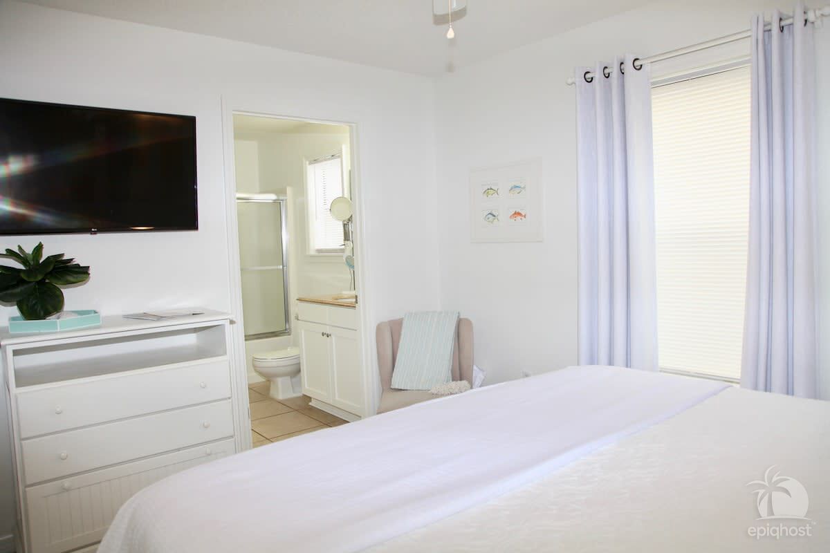 Un dormitorio con cama, televisión montada en la pared, cómoda y baño visible a través de una puerta abierta.