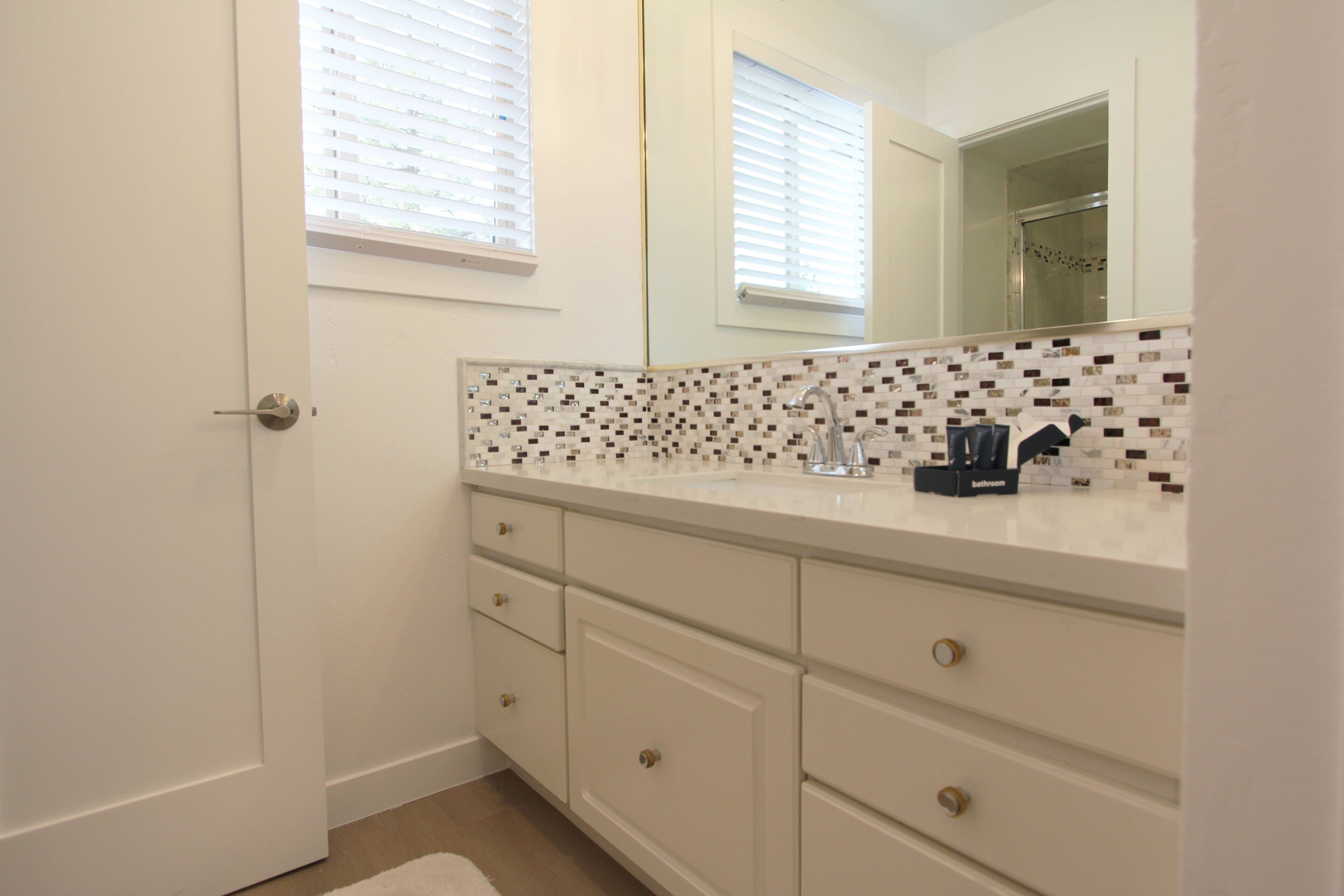 A bathroom with white cabinets, a tiled backsplash, and a shower visible in the mirror reflection.