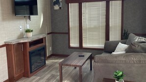 A modern living room with a flat-screen TV, a ceiling fan, a fireplace, a large window with blinds, and a sofa. - Great Getaway in adorable Cherokee Landing at Indian Lake with AC, WiFi (Lakeview)