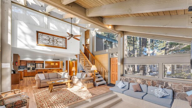 A spacious living area with a staircase, wooden flooring, and large windows offering a view of the outdoors. - Steps to Beach | Deck | Community Pool & More (South Lake Tahoe)